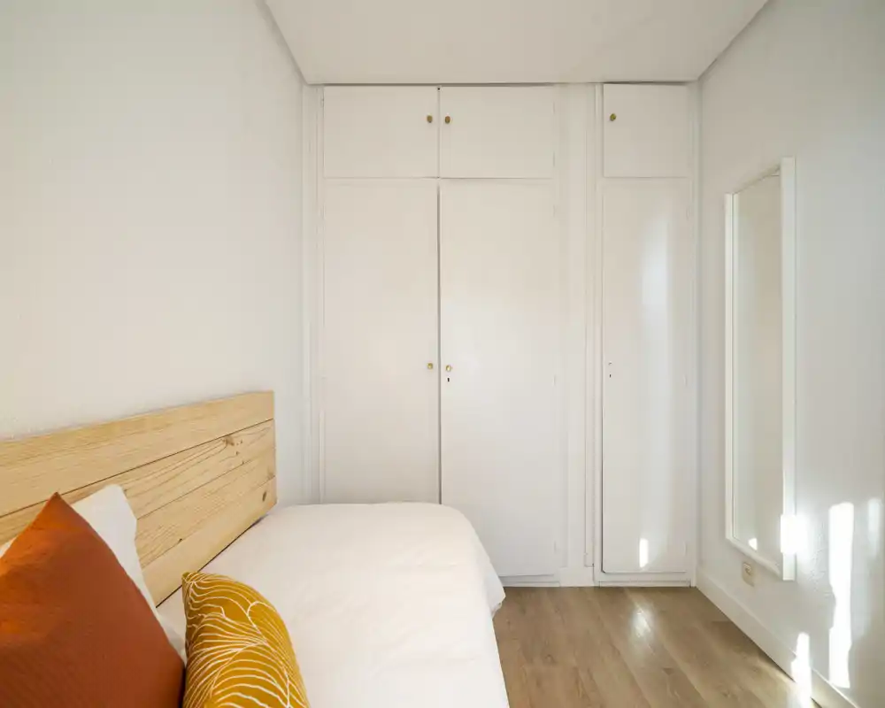 Camera da letto luminosa e ordinata con testiera in legno, biancheria bianca e cuscini accentuati. Armadi a muro e specchio a figura intera ottimizzano lo spazio e la luce.