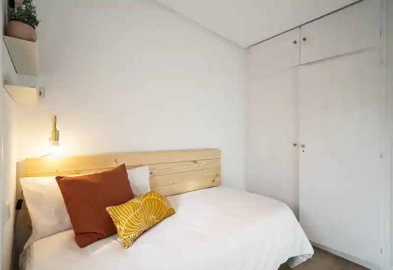 Camera da letto compatta e luminosa con testiera in legno, cuscini colorati e armadio a muro — ambiente accogliente e ben illuminato, ideale per un soggiorno riposante.