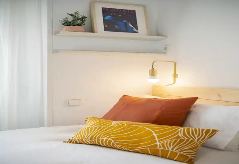 Bright, cozy bedroom corner with a neatly made bed, warm bedside lamp and decorative pillows. Clean white walls and soft natural light create a welcoming atmosphere ideal for listings.