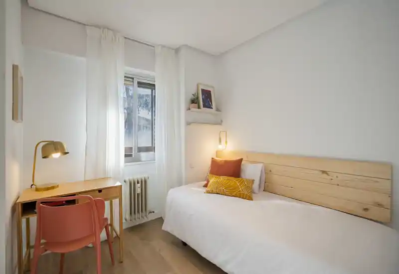 Camera singola luminosa e ben arredata con testata in legno, lampada da comodino calda, scrivania compatta e sedia vicino alla finestra — ideale per studenti o brevi soggiorni.