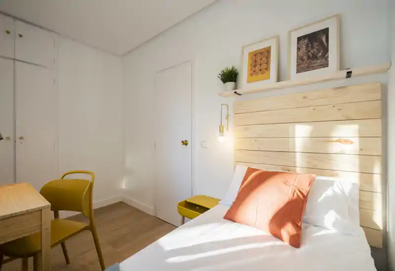 Camera singola moderna e luminosa con testiera in legno, biancheria bianca e un cuscino arancione. La luce naturale illumina la stanza; una scrivania e una sedia offrono un'area lavoro pratica.