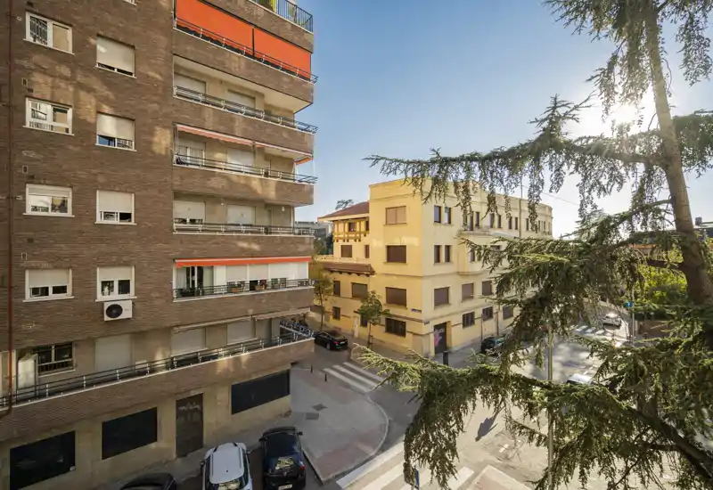 Vista soleada desde un balcón hacia edificios residenciales y un cruce con árboles, coches aparcados abajo y cielo azul despejado.