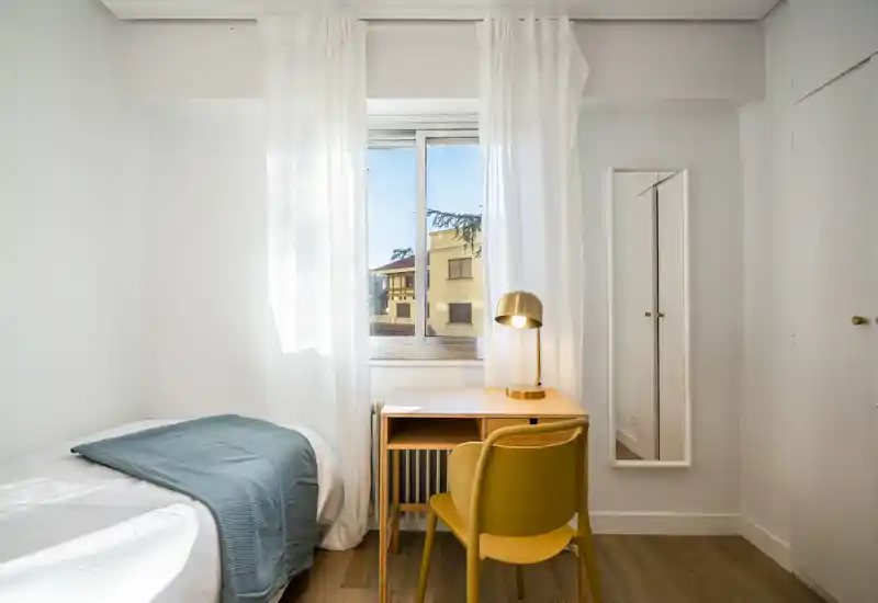 Dormitorio pequeño y luminoso con cama individual, escritorio junto a la ventana y una lámpara cálida — limpio, minimalista y bien iluminado para una estancia cómoda.
