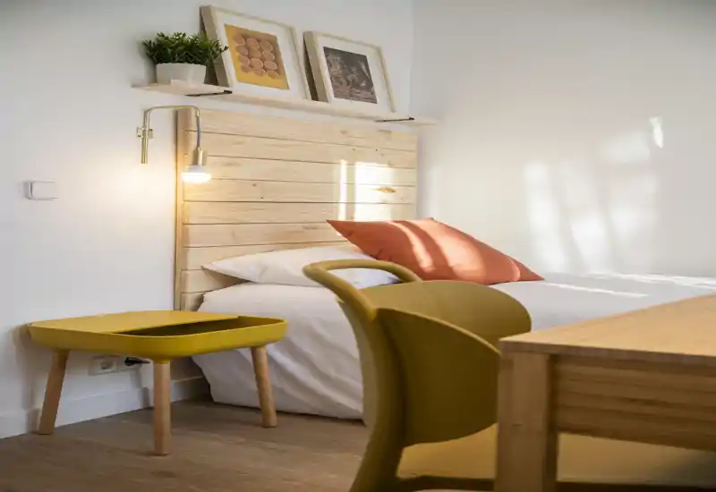 Camera da letto luminosa e moderna con testiera in legno, biancheria bianca e cuscino tono caldo. La stanza è ben illuminata e arredata, perfetta per un annuncio immobiliare che valorizza comfort e luce naturale.