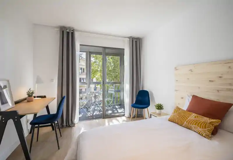 Camera da letto moderna e luminosa con testiera in legno, biancheria bianca e cuscini decorativi; grandi porte finestra si aprono su un balcone che illumina l'ambiente.