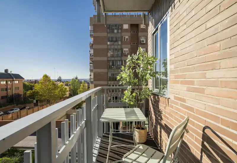 Balcón urbano soleado con una pequeña mesa y dos sillas, planta en maceta y una agradable vista de los edificios y árboles cercanos — ideal para el café matutino.