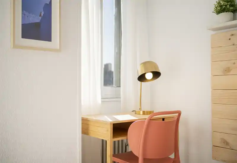 Pequeño rincón de trabajo luminoso con escritorio de madera, silla color coral y lámpara dorada junto a la ventana, ideal para concentrarse o estudiar.