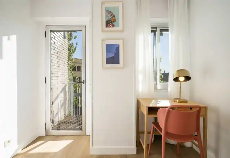 Pequeño acceso luminoso o rincón de estudio junto a una puerta acristalada que da a un balcón estrecho; escritorio de madera, silla color coral y lámpara dorada crean un acogedor espacio de trabajo.