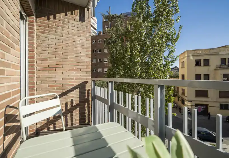 Pequeño balcón soleado con barandilla de metal, silla plegable y mesa, con vistas a una calle arbolada — perfecto para el café de la mañana.