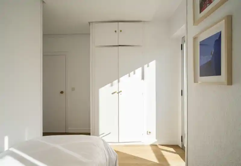 Rincón de dormitorio luminoso y minimalista con cama bien hecha, armario empotrado y suelo de madera cálida; la luz solar crea un ambiente acogedor.
