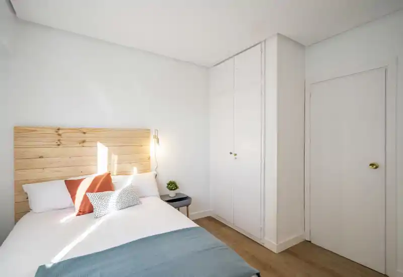 Bright, minimalist bedroom with a wooden headboard, white linens and decorative cushions; natural light and built-in wardrobe create a clean, inviting space.