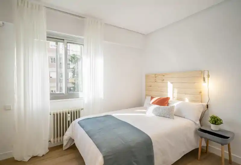 Bright, cozy bedroom with a wooden headboard, crisp white linens and decorative pillows. Natural light fills the room through a window with sheer curtains, creating a calm and inviting atmosphere.