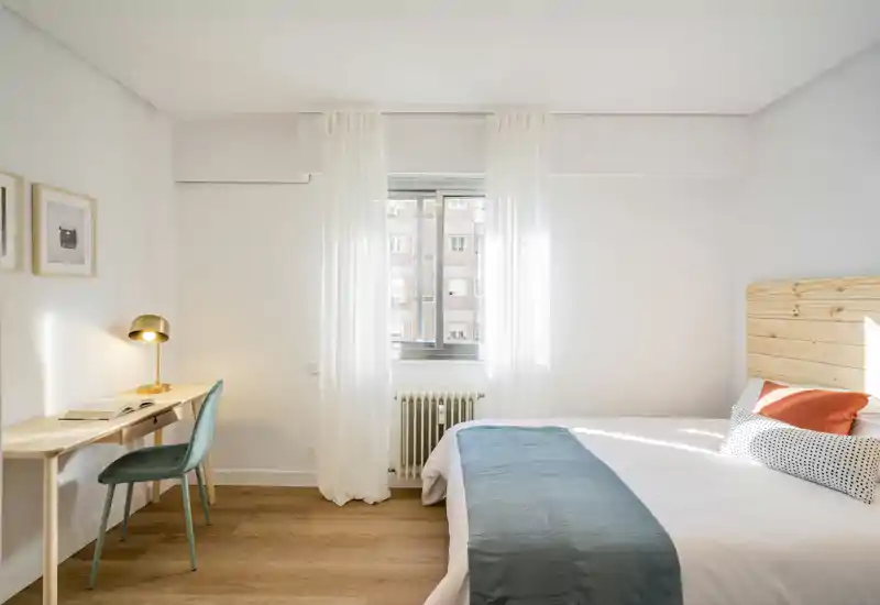 Bright, minimalist bedroom with a wooden headboard, neatly made double bed, desk and window letting in abundant natural light — ideal for short-term rentals.