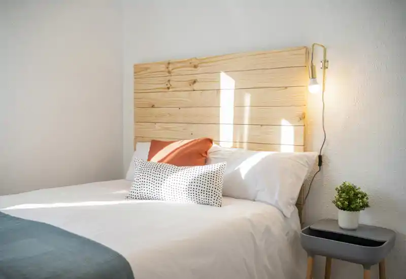 Sunny, neatly styled bedroom with a wooden headboard, white linens and decorative pillows; a small bedside table with a plant and a wall lamp create a warm, inviting atmosphere.