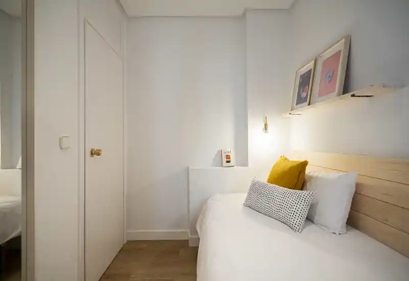 Dormitorio compacto y luminoso con cabecero de madera, ropa de cama blanca impecable y cojines decorativos. Una pequeña estantería con cuadros y una lámpara de pared cálida aportan un toque acogedor y moderno.