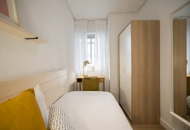 Dormitorio compacto y luminoso con una cama individual bien arreglada, cabecero de madera, escritorio junto a la ventana y armario. Estilo limpio de inspiración escandinava y luz natural agradable.