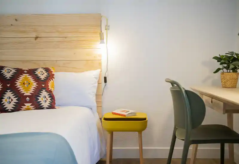 Coin chambre lumineux et soigné avec tête de lit en bois, coussin à motifs, linge de lit blanc et petite table de chevet jaune; un bureau et une chaise à droite complètent cet espace nuit cosy.