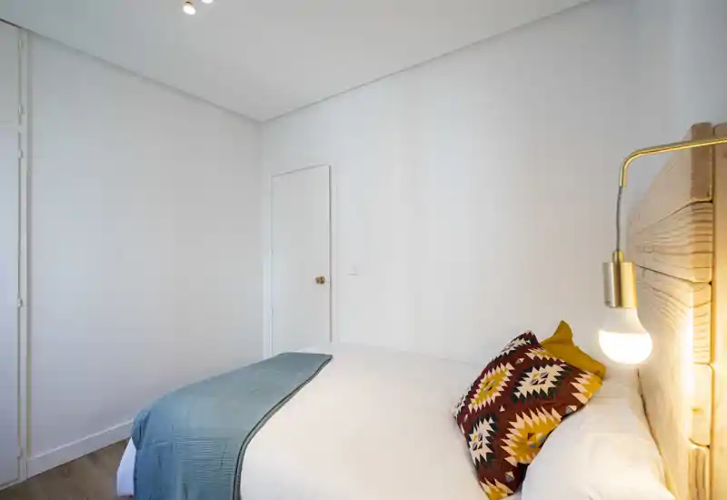 Camera da letto luminosa e accogliente con testiera in legno, cuscino decorativo e copriletto turchese; arredo minimale e illuminazione calda al comodino.
