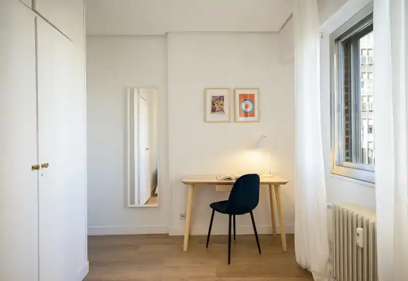 Rincón de dormitorio luminoso y minimalista con escritorio de madera, silla azul marino y armario; la luz natural y los cuadros aportan un espacio de trabajo tranquilo dentro de la habitación.