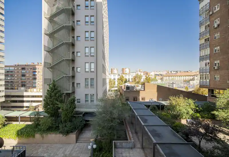Vista luminosa de un patio/terraza urbana con edificios residenciales alrededor, zonas verdes y pasarelas cubiertas — perfecta para mostrar las áreas exteriores comunes y el entorno del barrio.
