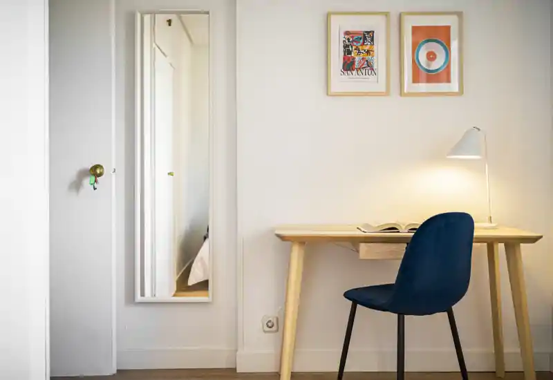 Rincón de dormitorio luminoso y minimalista con escritorio de madera y silla azul; un espejo refleja una pequeña parte de la cama. Cuadros y una lámpara de lectura añaden calidez.