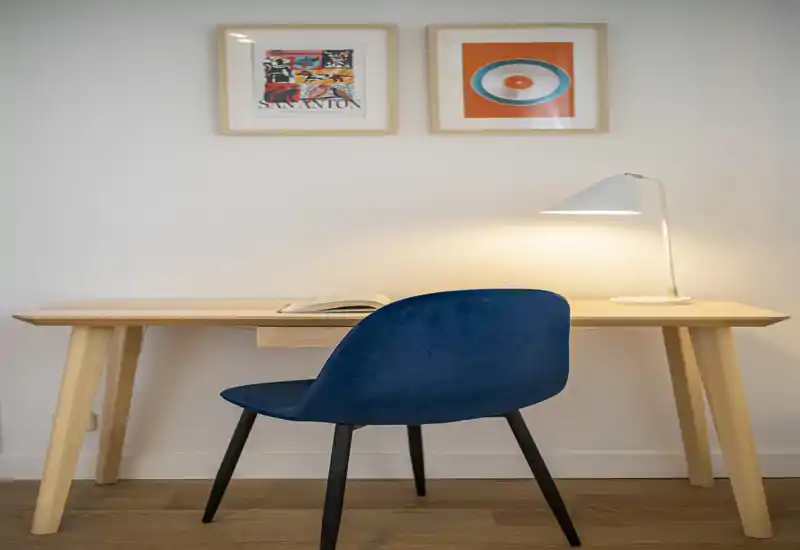 Espacio de trabajo compacto y moderno con escritorio de madera clara, silla azul y lámpara de mesa — rincón luminoso ideal para teletrabajo o lectura.