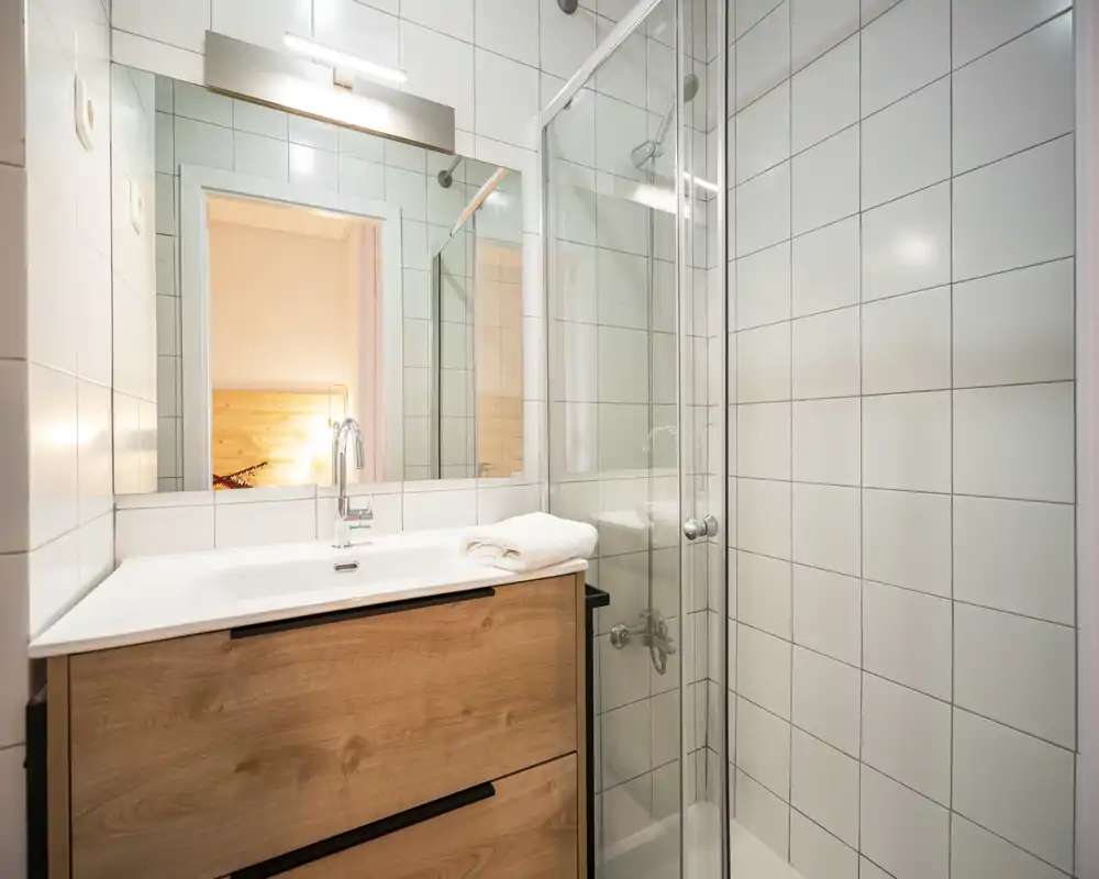 Bagno moderno e luminoso con piastrelle bianche, box doccia in vetro e mobile lavabo in legno con specchio.