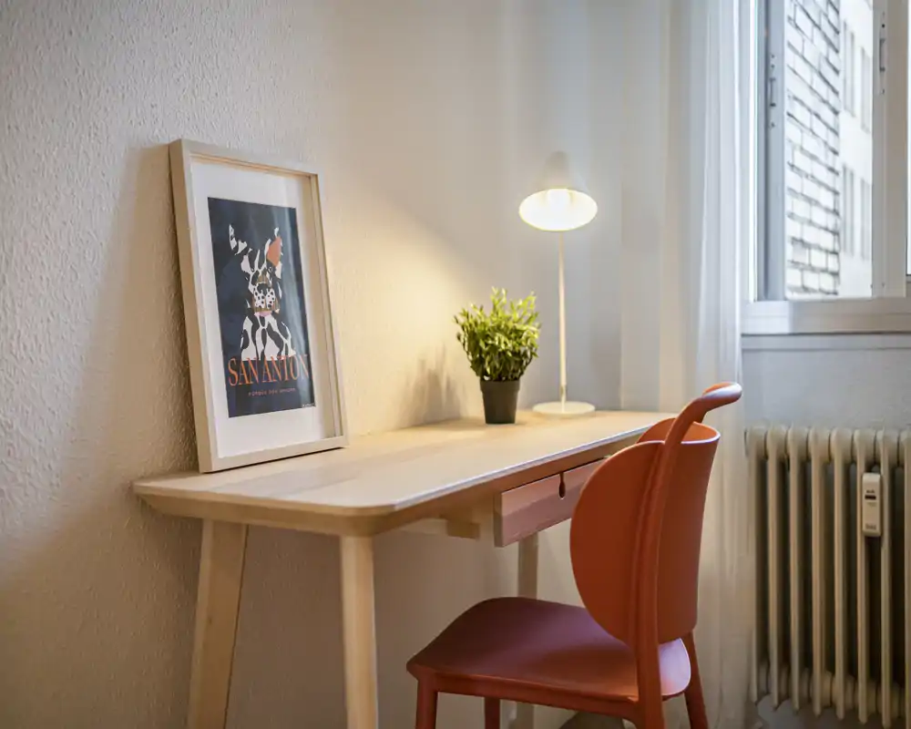 Angolo studio luminoso e curato con una scrivania in legno, lampada da tavolo, quadro incorniciato e sedia rossa accanto alla finestra e al radiatore.