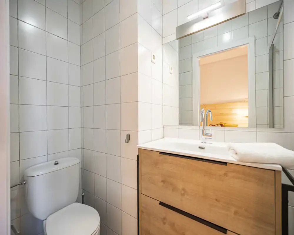Bagno moderno e pulito con pareti piastrellate bianche, WC compatto e mobile lavabo in legno con specchio, ben illuminato e ordinato.