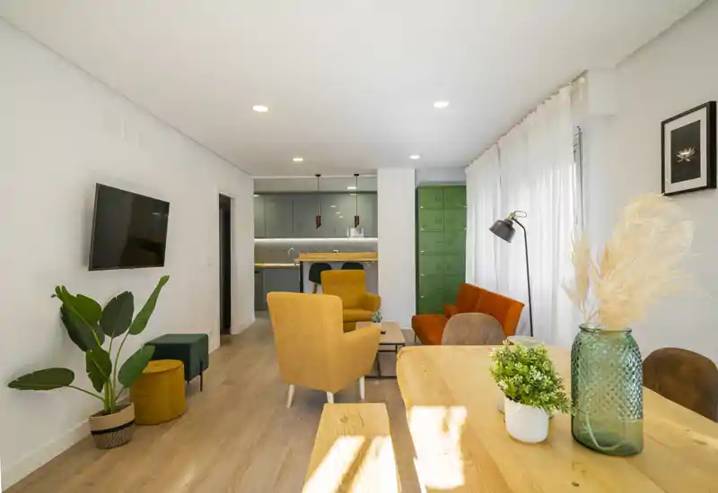 Salón moderno y luminoso con butacas de colores, televisor de pared y cocina abierta al fondo. La luz natural y las plantas crean un ambiente cálido y acogedor, ideal para un anuncio de alquiler.