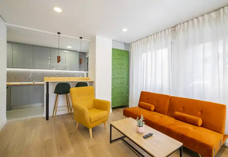 Zona de estar moderna y luminosa con sofá naranja llamativo, butaca mostaza y cocina abierta con barra — perfecta para alquileres urbanos con estilo.