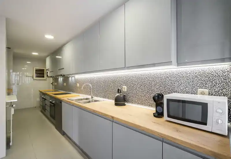 Cocina en galera elegante con armarios grises, encimeras de madera y electrodomésticos integrados; bien iluminada y moderna, con luz bajo los muebles y espacio práctico para cocinar.