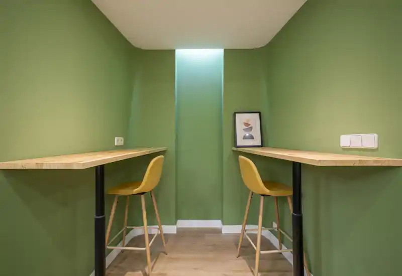 Pequeña zona tipo nicho con dos barras altas de madera y taburetes mostaza frente a paredes verdes; decoración minimal y luz suave en la parte superior crean un práctico espacio de trabajo o rincón para desayunar.