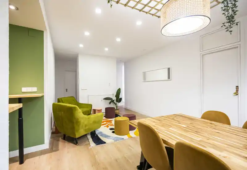 Zona de estar luminosa y decorada con mesa de comedor de madera en primer plano, butacas de terciopelo, alfombra colorida y pufs: un espacio abierto y acogedor para salón y comedor.
