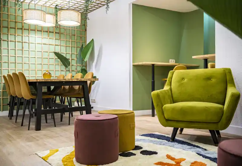 Área de estar luminosa y moderna con un sillón verde, pufs coloridos y una mesa de comedor al fondo. La iluminación cálida y la alfombra texturizada crean un ambiente acogedor, perfecto para relajarse o reuniones íntimas.