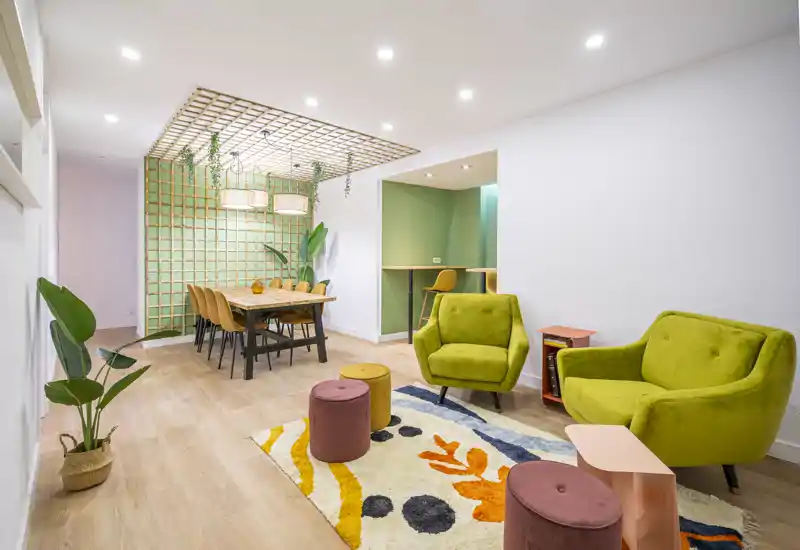 Zona de estar abierta y luminosa con sillones coloridos, alfombra estampada y mesa de comedor bajo una celosía decorativa en el techo — espacio moderno y acogedor ideal para reunirse.