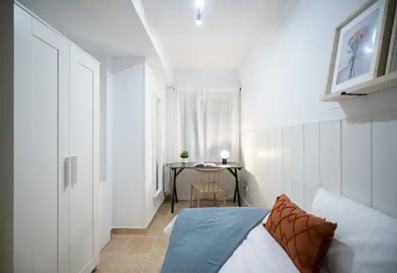 Camera compatta e ben illuminata con letto ordinato, cuscino decorativo e una piccola scrivania vicino alla finestra — ideale per una persona o soggiorni brevi.