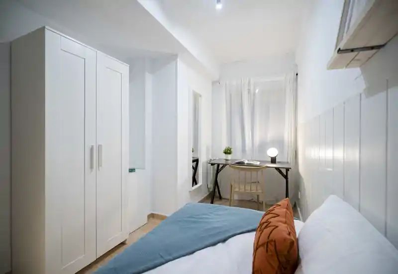 Camera da letto luminosa e minimal con letto in primo piano, scrivania vicino alla finestra e armadio — ideale per un singolo o soggiorni brevi.