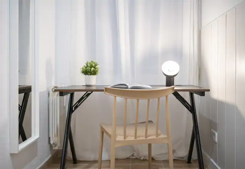 Small bright workspace with a minimalist desk, chair, bedside lamp and a potted plant in front of sheer curtains — ideal for focused work.