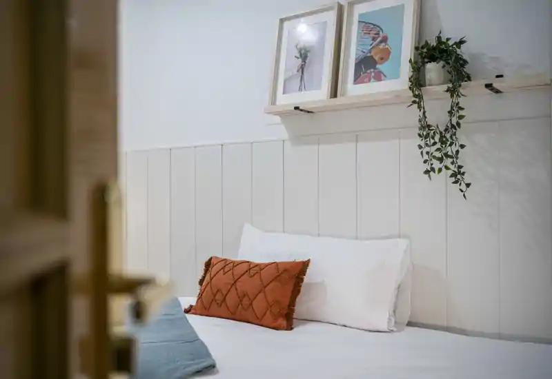 Bright, styled bedroom with a neatly made bed, decorative cushions and a small shelf with framed prints and a hanging plant — ideal for cozy rental photos.