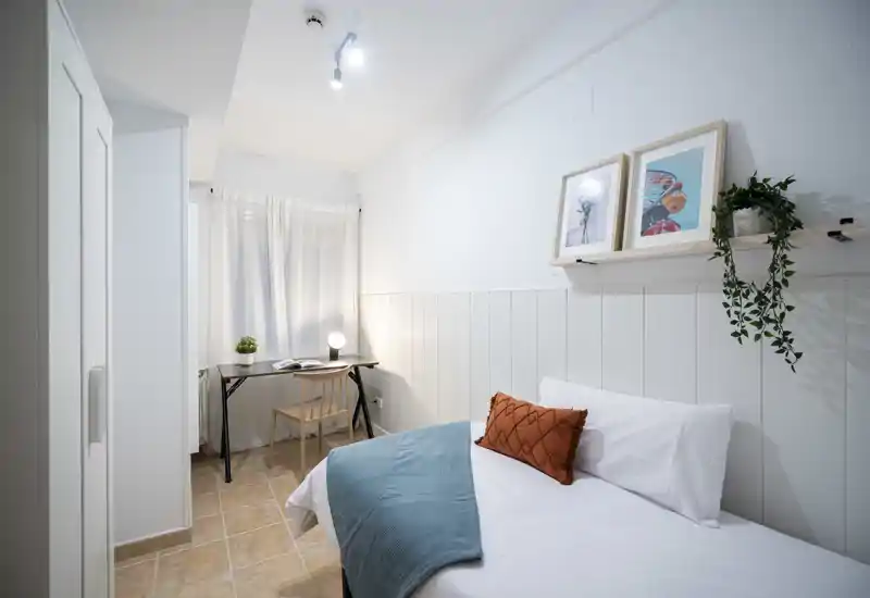 Camera da letto luminosa e ordinata con un letto singolo confortevole, tessili accoglienti, una scrivania semplice vicino alla finestra e quadri di gusto — ideale per uno studente o viaggiatore singolo.