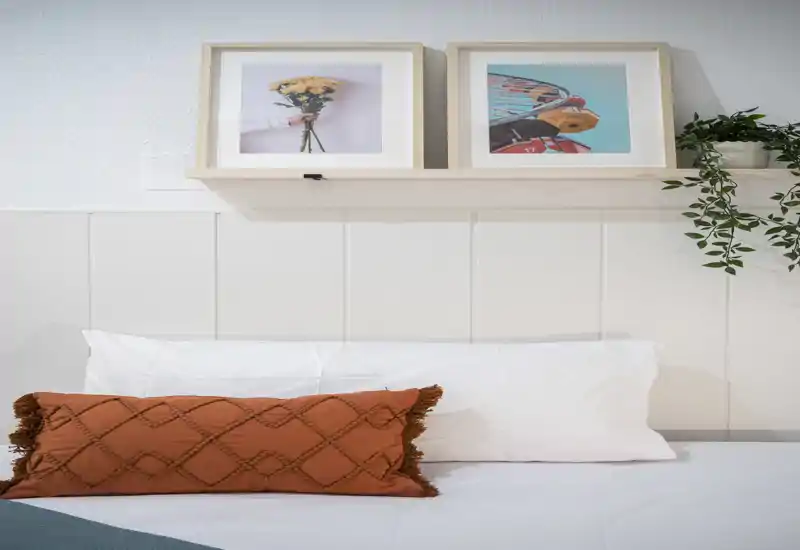 Camera da letto luminosa e curata, primi piani di cuscini bianchi e un cuscino decorativo color ruggine con quadri su una mensola — un angolo riposo accogliente, perfetto per gli annunci.