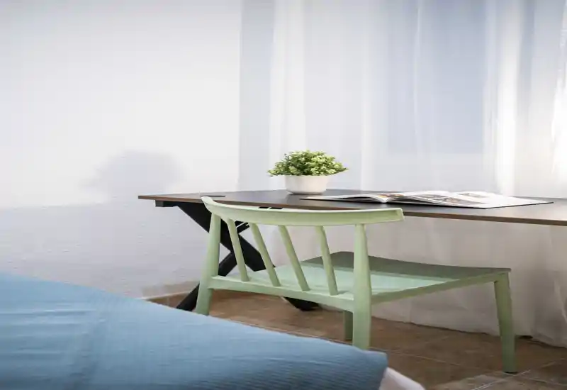 Rincón de dormitorio luminoso y de estilo sencillo con la cama parcialmente visible en primer plano, un escritorio junto a la ventana y un banco/silla verde menta con una pequeña planta.