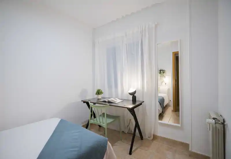 Chambre lumineuse et chaleureuse avec un lit confortable, un petit bureau et une décoration soignée. Les rideaux et le miroir pleine hauteur apportent de la profondeur.