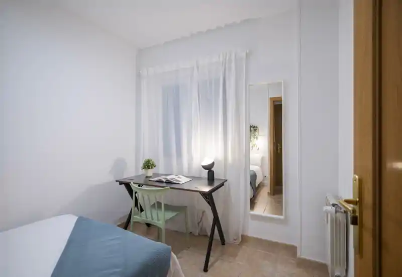 Dormitorio luminoso y bien presentado con cama visible, un escritorio estrecho y una silla verde junto a la ventana; paredes neutras y cortinas traslúcidas que crean un ambiente tranquilo.