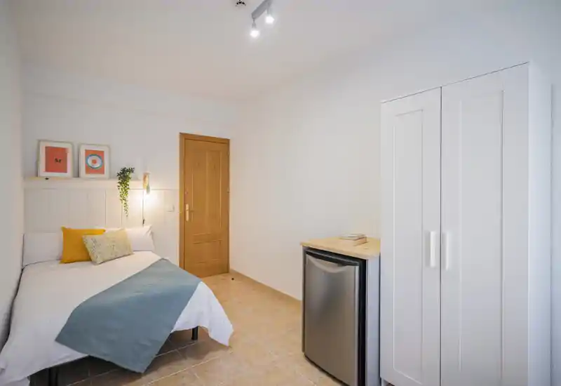 Habitación individual limpia y luminosa con cama cómoda, cojines decorativos y un armario compacto. Detalles en madera y suelo de baldosa, ideal para una estancia acogedora en solitario.