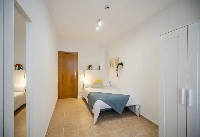 Bright, neatly styled single bedroom with a comfortable bed, decorative pillows and a teal throw; white walls and warm floor tiles create a clean, welcoming space.