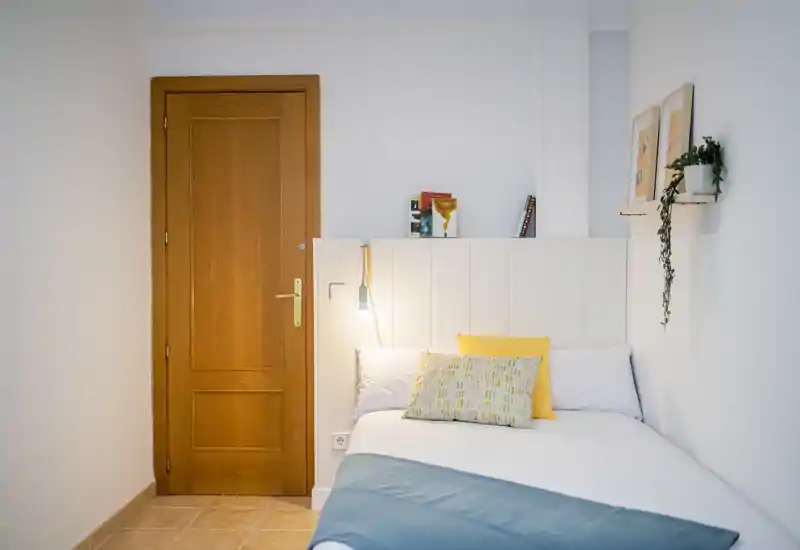 Compact, bright bedroom with a neatly made single bed, headboard-mounted reading lamp and soft decorative cushions. The space features warm wood accents and minimal, modern decor for a cozy, functional stay.