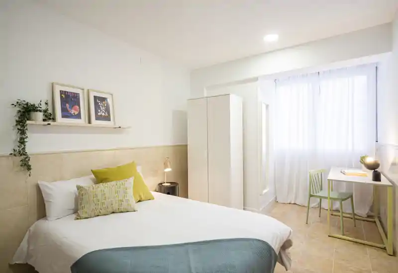 Dormitorio luminoso y bien decorado con cama doble, cojines, armario y un pequeño escritorio junto a la ventana, perfecto para descansar o trabajar.