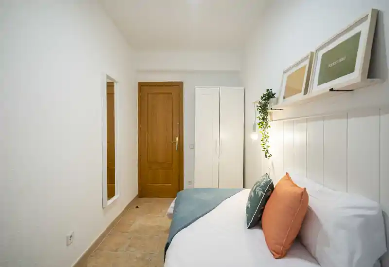 Camera da letto luminosa e compatta con letto matrimoniale in lenzuola bianche, cuscini colorati e plaid verde acqua; stampe incorniciate e una pianta appesa donano un tocco accogliente.